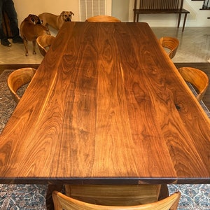 May include: A large, rectangular wooden dining table with a rich, dark brown finish. The table is surrounded by several wooden chairs with curved backs. The wood grain is prominent, and the table appears to be handcrafted. The overall style is modern and rustic.