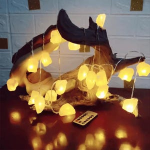 Puede incluir: Una escultura decorativa de madera con luces de cadena LED blanco cálido. Las luces tienen forma de pequeñas piedras irregulares. Un pequeño mando a distancia es visible en la superficie inferior.