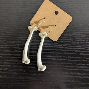May include: A pair of bone-shaped earrings with gold-colored hooks. The earrings are white and have a curved shape, resembling small bones. They are displayed on a brown cardboard earring display card.