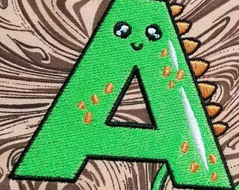 May include: Embroidered green letter "A" with a dinosaur design. The letter features a smiling face, orange spikes, and decorative accents. The background has a brown and tan swirled pattern.