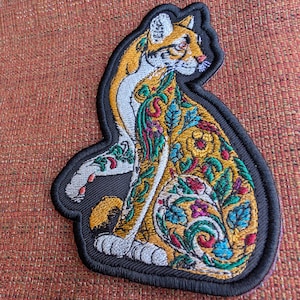 May include: An embroidered patch featuring a seated cat with a golden yellow coat, white chest, and paws. The cat's body is decorated with intricate, colorful floral and leafy patterns in shades of green, red, blue, and yellow, all outlined in black.