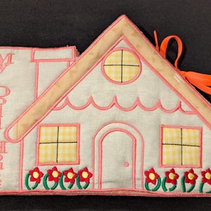May include: A fabric dollhouse book with a house-shaped cover. The cover is white with pink trim, a beige roof, and yellow gingham windows. The words "My Doll House" are embroidered on the side. Red flowers with green stems decorate the bottom.