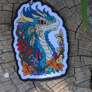 May include: An embroidered patch featuring a blue and white dragon head surrounded by colorful autumn leaves, small purple flowers, and clusters of red berries. The design is set against a white background with a black border and a metal loop at the top.