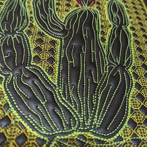Puede incluir: Primer plano de un artículo de cuero negro con un diseño de cactus bordado detallado. El cactus está delineado con hilo verde brillante, con una flor roja en la parte superior. El fondo presenta un patrón geométrico amarillo y dorado.