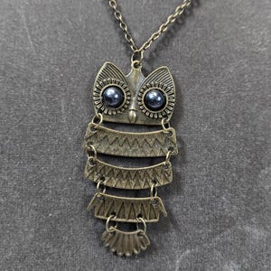May include: Antique bronze owl pendant necklace. The owl pendant features a detailed design with layered sections and dark blue bead eyes. The necklace chain is also bronze-toned, creating a cohesive and vintage aesthetic.