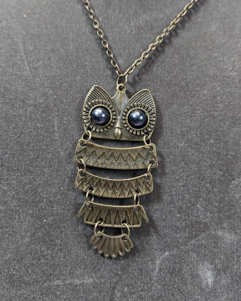 May include: Antique bronze owl pendant necklace. The owl pendant features a detailed design with layered sections and dark blue bead eyes. The necklace chain is also bronze-toned, creating a cohesive and vintage aesthetic.