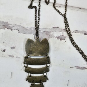 May include: A bronze-colored pendant necklace with a long chain. The pendant has an owl-like shape with layered, rectangular and scalloped elements. The necklace is displayed against a white, distressed wood background.