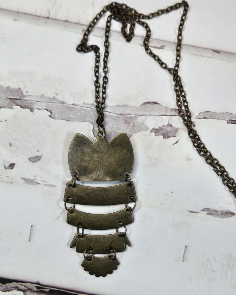 May include: A bronze-colored pendant necklace with a long chain. The pendant has an owl-like shape with layered, rectangular and scalloped elements. The necklace is displayed against a white, distressed wood background.