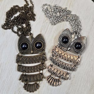 May include: Two owl pendant necklaces. One is bronze-toned with a matching chain, the other is silver-toned with a silver chain. Both owl pendants have black bead eyes and layered, geometric body designs.