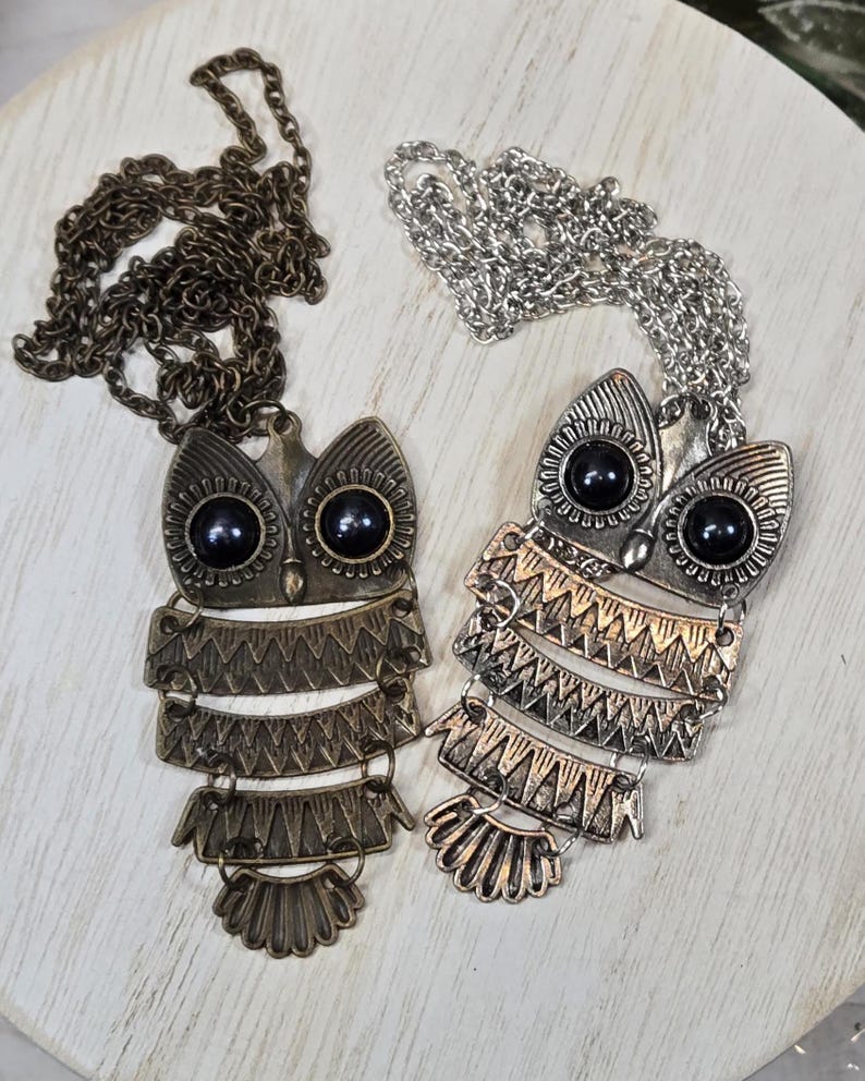 May include: Two owl pendant necklaces. One is bronze-toned with a matching chain, the other is silver-toned with a silver chain. Both owl pendants have black bead eyes and layered, geometric body designs.