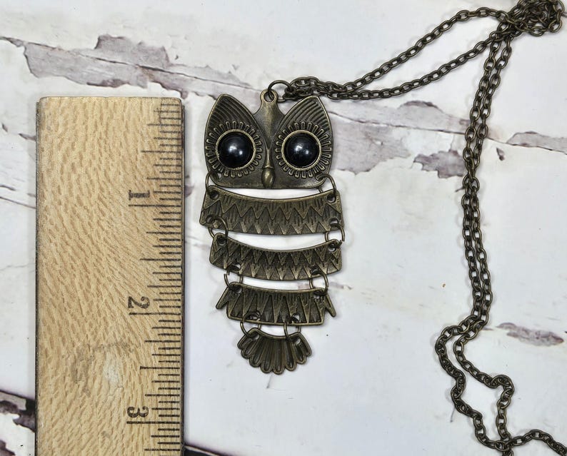 May include: A bronze-toned owl pendant necklace. The owl has black bead eyes and layered, decorative feathers. The chain is also bronze-toned. The pendant measures approximately 3 inches in length.