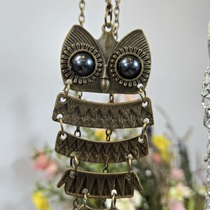 May include: A bronze-toned owl pendant necklace. The owl design features a detailed face with dark bead eyes and layered, textured body segments. The necklace hangs from a delicate chain, showcasing an antique aesthetic.
