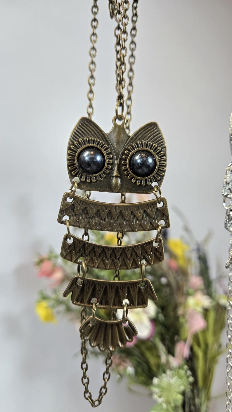 May include: A bronze-toned owl pendant necklace. The owl design features a detailed face with dark bead eyes and layered, textured body segments. The necklace hangs from a delicate chain, showcasing an antique aesthetic.