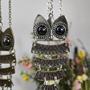 May include: Silver-toned owl pendant necklace with black bead eyes and layered, patterned metalwork. The owl pendant hangs from a delicate chain. The necklace is displayed against a soft, blurred background with floral accents.