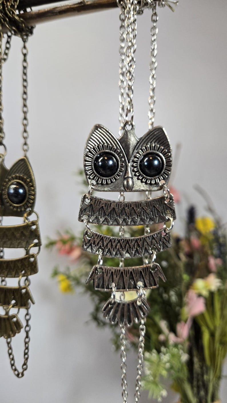 May include: Silver-toned owl pendant necklace with black bead eyes and layered, patterned metalwork. The owl pendant hangs from a delicate chain. The necklace is displayed against a soft, blurred background with floral accents.