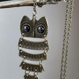 May include: A bronze-toned owl pendant necklace. The owl has large, dark eyes and a layered body design. The necklace chain is also bronze-toned and hangs from a branch. The pendant is a unique, handcrafted piece of jewelry.