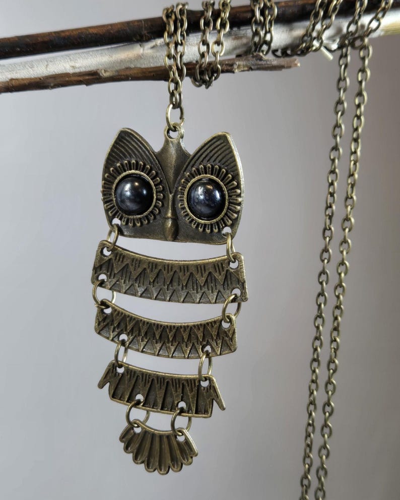 May include: A bronze-toned owl pendant necklace. The owl has large, dark eyes and a layered body design. The necklace chain is also bronze-toned and hangs from a branch. The pendant is a unique, handcrafted piece of jewelry.