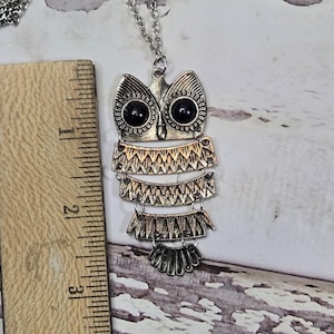 May include: Silver-toned owl pendant necklace. The owl pendant has black eyes and layered, textured details. The necklace is approximately 3 inches long, as measured by the wooden ruler in the image. The chain is silver and delicate.