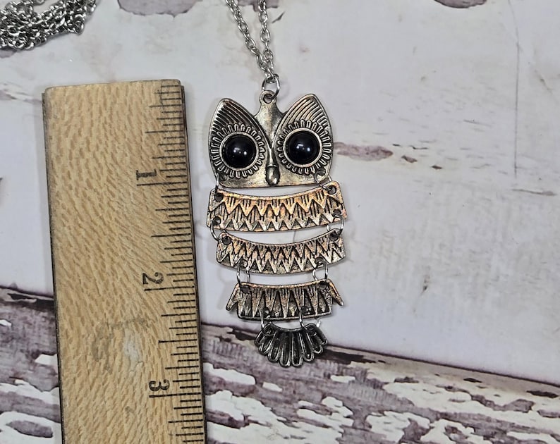 May include: Silver-toned owl pendant necklace. The owl pendant has black eyes and layered, textured details. The necklace is approximately 3 inches long, as measured by the wooden ruler in the image. The chain is silver and delicate.