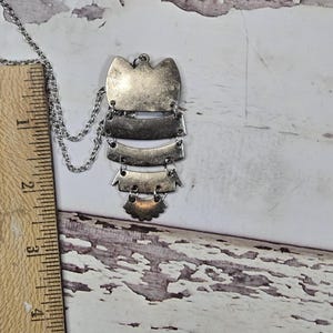 May include: Silver-toned pendant necklace with a layered design. The pendant features a series of rectangular and curved shapes, suspended from a delicate chain. The necklace is displayed against a weathered, white wooden surface.