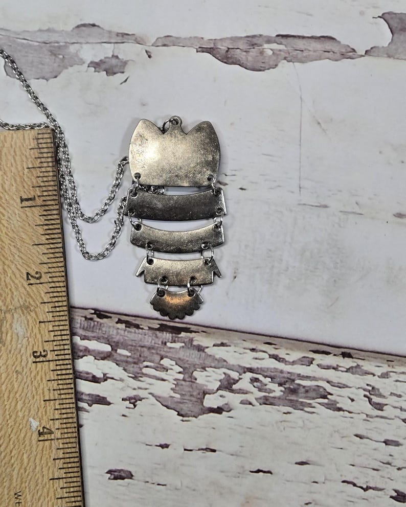 May include: Silver-toned pendant necklace with a layered design. The pendant features a series of rectangular and curved shapes, suspended from a delicate chain. The necklace is displayed against a weathered, white wooden surface.