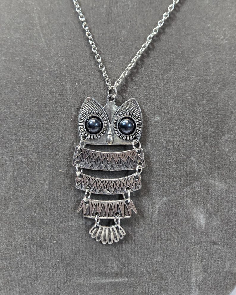 May include: Silver-toned owl pendant necklace. The owl pendant features a layered design with intricate detailing and dark gray eyes. The necklace has a delicate chain and a simple clasp.