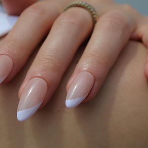 Könnte beinhalten: Nahaufnahme einer Hand mit vier ovalen Nägeln, die mit einer klassischen French Manicure lackiert sind. Die Nägel sind hellrosa mit einer weißen Spitze.