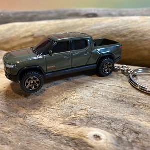 May include: A green Rivian R1T pickup truck keychain. The truck is a toy model with black wheels and a silver key ring.