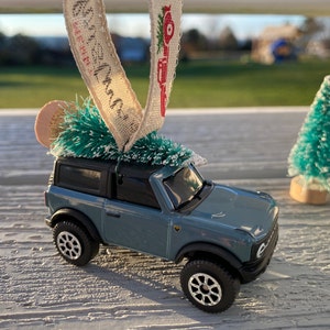 May include: A blue toy SUV with a Christmas tree on top, decorated with a small straw hat and a ribbon with text. The SUV has black tires and a black roof. Miniature green Christmas trees are in the background.