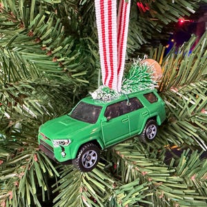 May include: A green toy SUV vehicle ornament featuring a small artificial pine tree secured to the roof with a red and white striped fabric ribbon loop for hanging. The miniature car has black wheels, dark windows, and detailed front headlights.