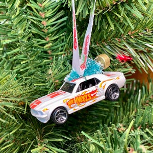 May include: A white Hot Wheels car ornament with red and orange flame details and the Hot Wheels logo. The car is suspended by a white ribbon and sits among green Christmas tree needles.