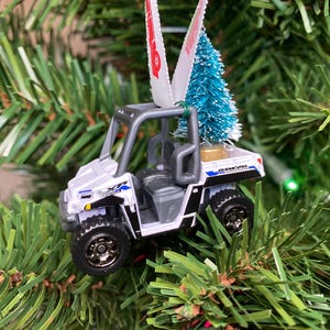 May include: White toy utility vehicle with a small blue Christmas tree in the bed. The vehicle is a Christmas ornament.