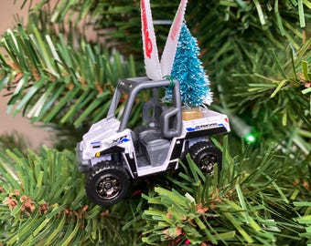 May include: White toy utility vehicle with a small blue Christmas tree in the bed. The vehicle is a Christmas ornament.