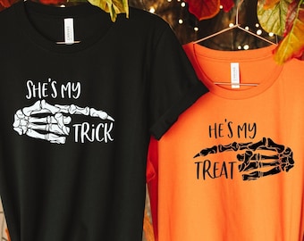 May include: Two t-shirts, one black and one orange, hanging on a clothes rack. The black shirt has the text "She's my Trick" with a skeleton hand graphic. The orange shirt says "He's my Treat" with a matching skeleton hand.