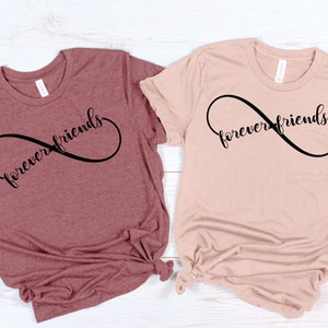 May include: Two t-shirts with the text "forever friends" written in a cursive font inside an infinity symbol. The t-shirt on the left is maroon and the t-shirt on the right is light pink.