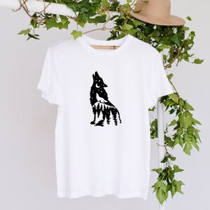 May include: White t-shirt with a black wolf silhouette howling at the moon, set against a forest and starry sky. The design is on a wooden hanger. The shirt is a simple, casual garment.