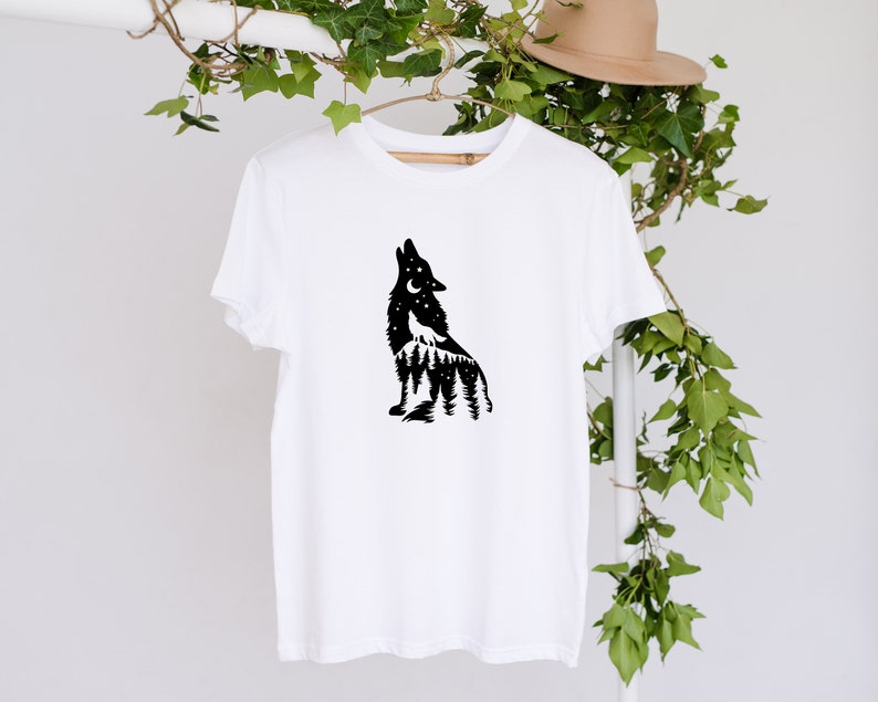 May include: White t-shirt with a black wolf silhouette howling at the moon, set against a forest and starry sky. The design is on a wooden hanger. The shirt is a simple, casual garment.