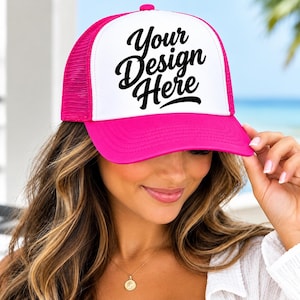 May include: A bright pink and white trucker hat featuring a foam front panel with the black text Your Design Here printed in a cursive font. The cap includes a solid pink brim and a breathable mesh back panel with an adjustable snapback closure.