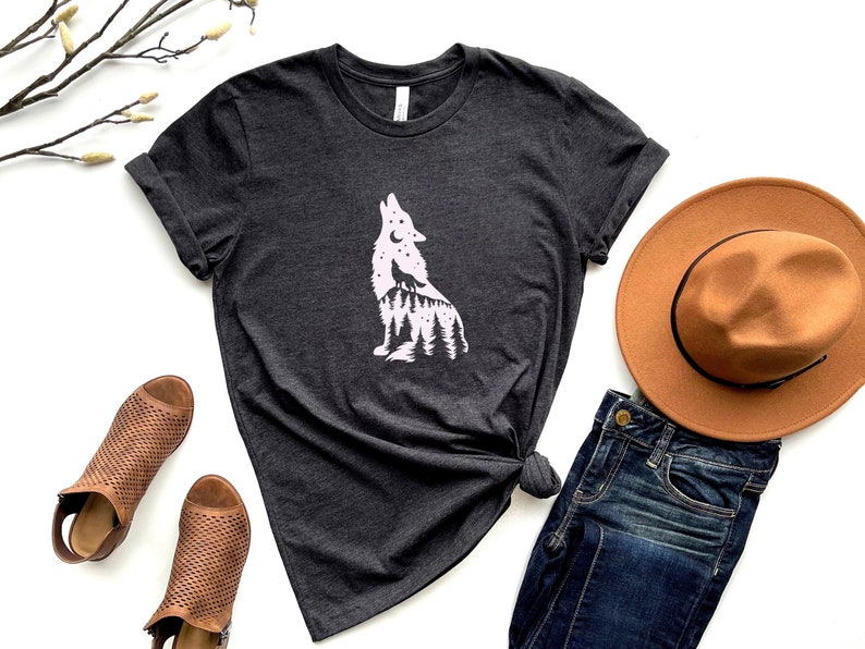 May include: Charcoal grey t-shirt with a white wolf silhouette graphic, incorporating a forest and moon. The image includes a brown hat, blue jeans, and brown open-toe shoes. The t-shirt is knotted at the waist for styling.