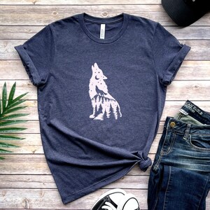 May include: A navy blue t-shirt with a white wolf silhouette design, incorporating a forest and stars. The shirt is displayed with blue jeans, a black cap, and trainers on a wooden surface.