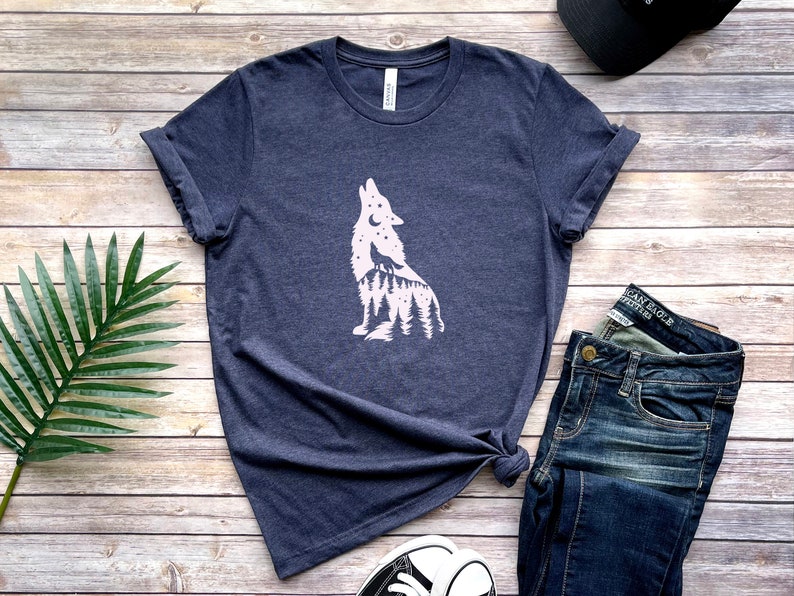 May include: A navy blue t-shirt with a white wolf silhouette design, incorporating a forest and stars. The shirt is displayed with blue jeans, a black cap, and trainers on a wooden surface.