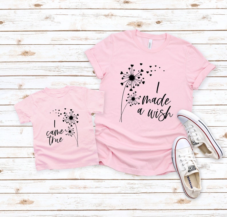 May include: Two pink t-shirts with black dandelion designs and the phrases "I made a wish" and "I came true." The shirts are next to a pair of white canvas sneakers with red and blue accents. The shirts are on a light wood background.