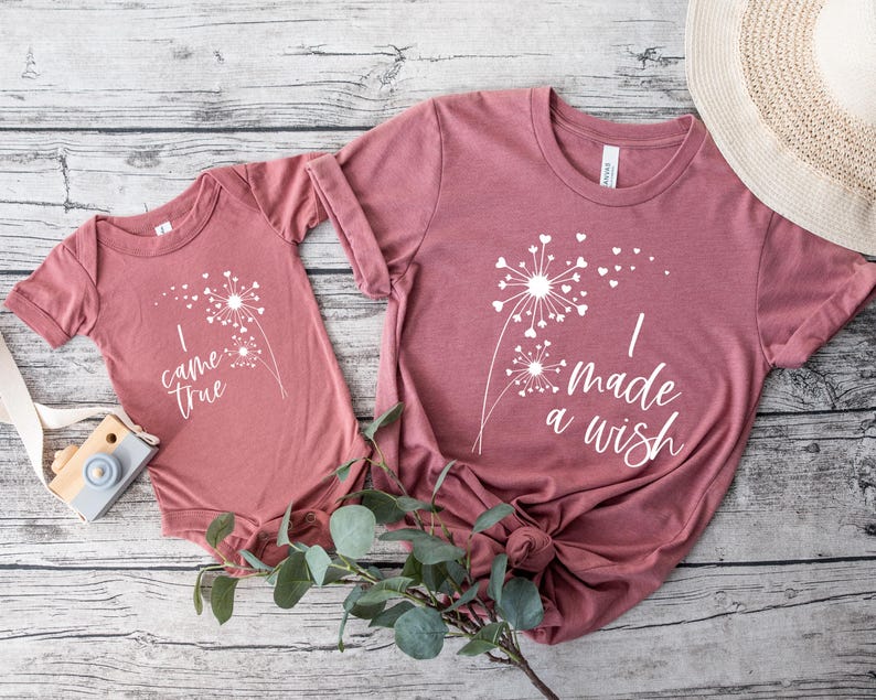May include: Matching mauve-colored t-shirts and a onesie with white dandelion graphics. The adult shirt reads "I made a wish," while the onesie says "I came true." A wooden toy camera and a straw hat are also in the image.