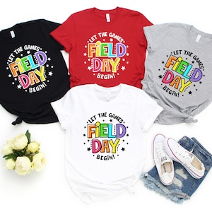 May include: Four t-shirts with the text "Let the games begin! Field Day" in a rainbow color scheme and surrounded by stars. The shirts are in black, red, grey, and white.