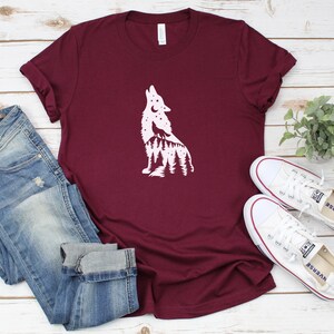 May include: A maroon t-shirt featuring a white wolf howling design, alongside blue jeans and white trainers. The wolf graphic includes a forest and moon. The shirt is folded, and the jeans are cuffed at the ankles.