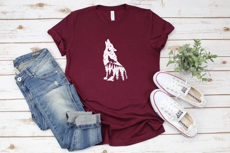 May include: A maroon t-shirt featuring a white wolf howling design, alongside blue jeans and white trainers. The wolf graphic includes a forest and moon. The shirt is folded, and the jeans are cuffed at the ankles.
