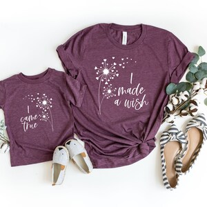 May include: Two maroon t-shirts, one adult and one child size, with white dandelion designs and text. The adult shirt says "I made a wish" and the child's says "I came true". The image includes shoes and greenery.