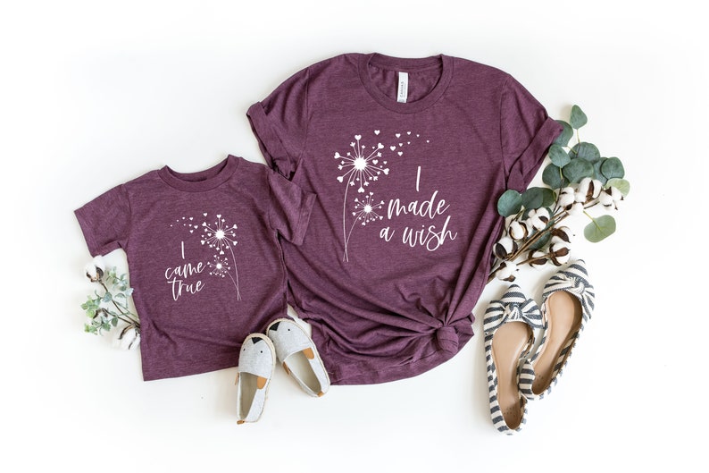 May include: Two maroon t-shirts, one adult and one child size, with white dandelion designs and text. The adult shirt says "I made a wish" and the child's says "I came true". The image includes shoes and greenery.