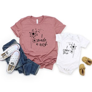 May include: A dusty rose t-shirt and a white onesie with matching dandelion designs and the phrases "I made a wish" and "I came true". The image also includes blue jeans, white sneakers, and baby moccasins.