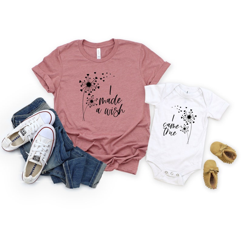 May include: A dusty rose t-shirt and a white onesie with matching dandelion designs and the phrases "I made a wish" and "I came true". The image also includes blue jeans, white sneakers, and baby moccasins.